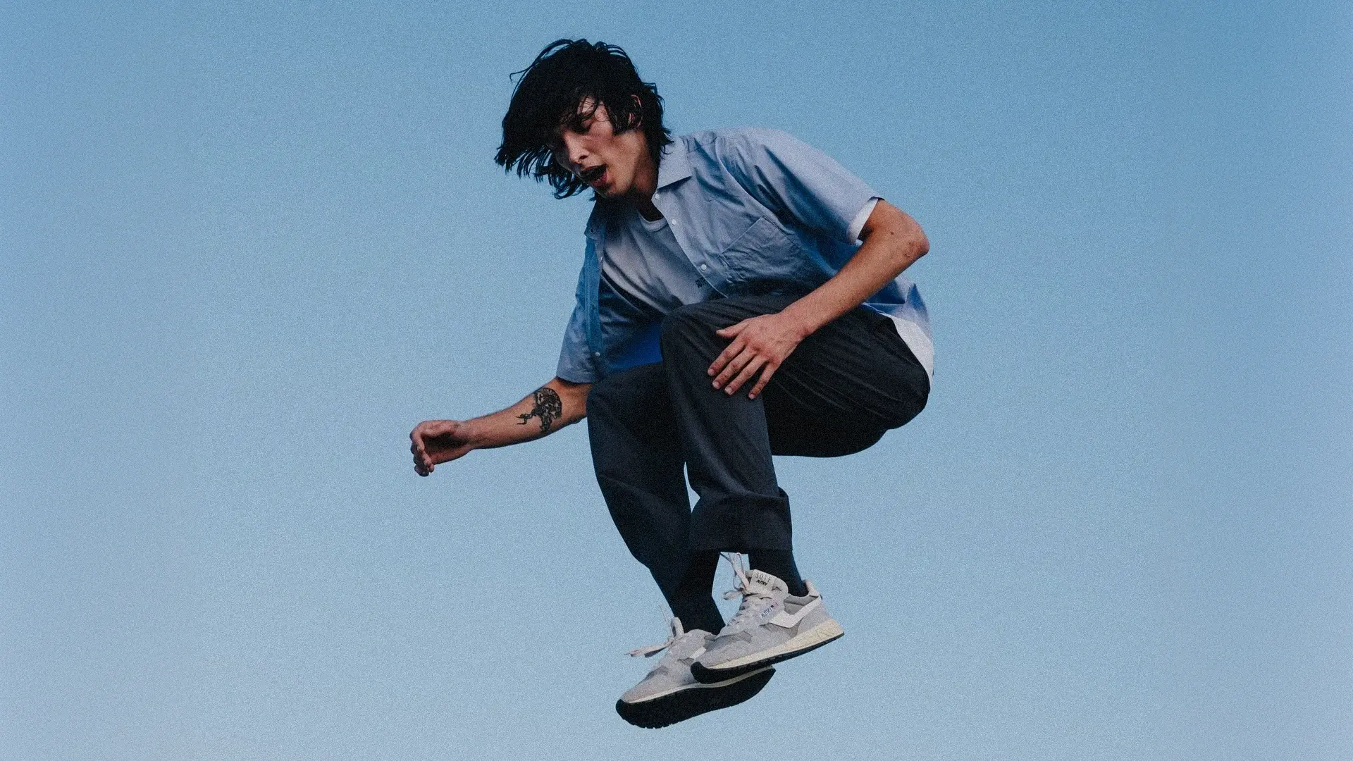 Young man with black hair wearing a light blue shirt and black pants is jumping against a clear blue sky.