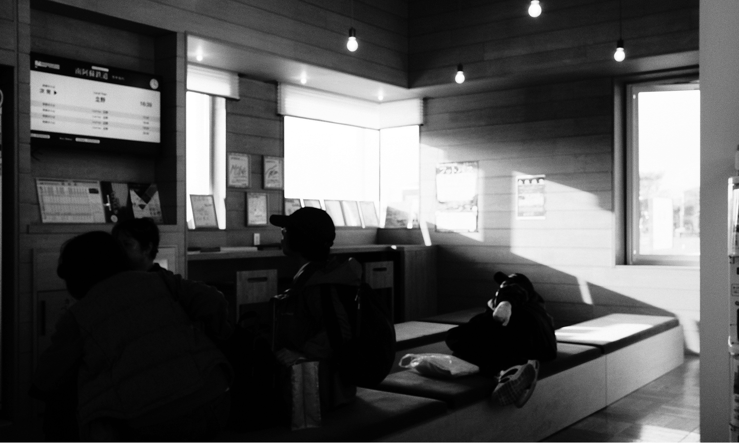 People sitting and resting in a waiting area with wooden panel walls and a digital information screen.